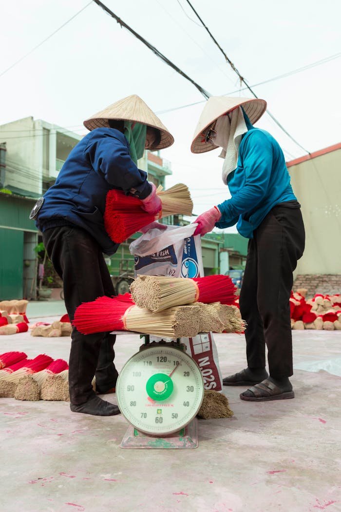 Home Two workers handling bundles of goods on a scale in an outdoor market setting.
